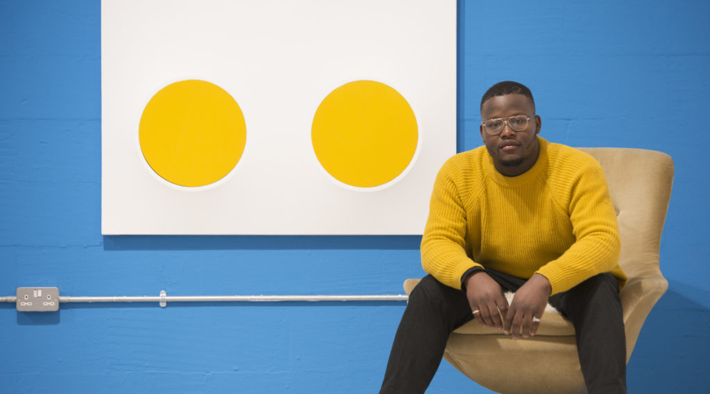 Jasper sat on a chair wearing a striking mustard jumper next to one of his works